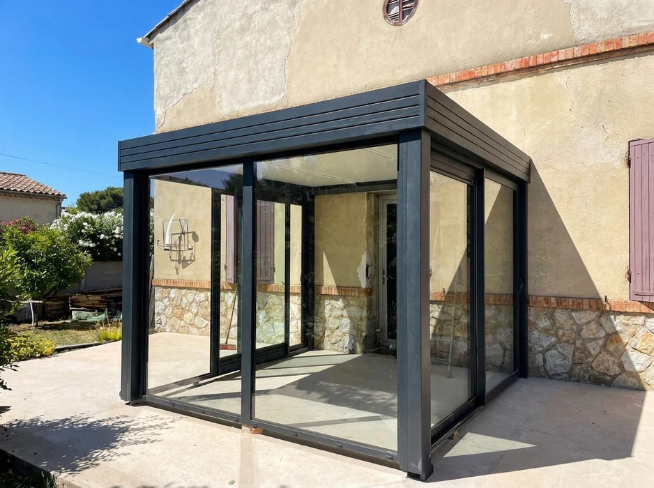 Véranda aluminium gris anthracite adossée à une maison en pierre, toiture plate à lames, parois entièrement vitrées avec baies coulissantes